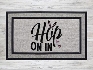 Mockup of an Easter themed doormat that features the phrase "Hop on In" with a cute pair of bunny ears over the 'O' in "Hop'