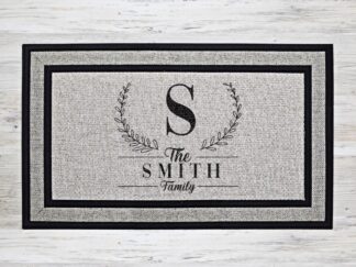 Mock up of a custom welcome doormat that features your last name's first initial as a monogram in a laurel wreath, with your family name underneath.