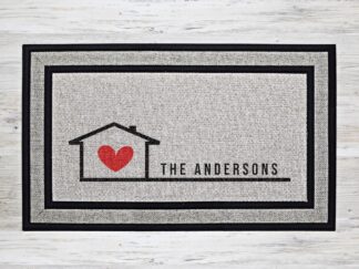 Mockup of a custom welcome doormat that features your last name in next to an outline of a house with a red heart in it, symbolizing home is where the heart is