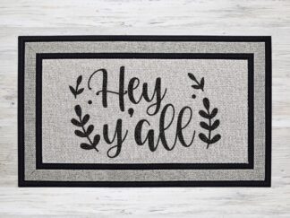 Mockup of a doormat that says, "Hey Y'all" in a bold black handwriting style font with leafy flourishes on both sides of the text.