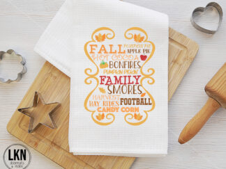 White dish towel with fall-themed phrases: pumpkin pie, apple pie, hot cocoa, bonfires, pumpkin pickin', family, smores, harvest, hayrides, football, candy corn.
