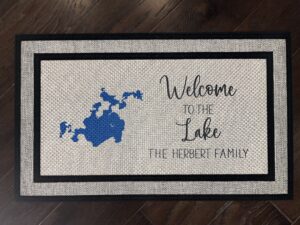 Photo of a doormat for a client at Woman Lake in Minnesota, for The Herbert Family, that says "Welcome to the Lake, The Herbert Family" with a cartography map of Woman Lake.