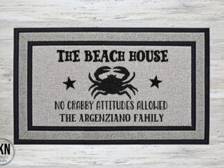 Mockup of a beach themed doormat that says "The Beach House - No Crabby Attitudes Allowed" in a bold black font with a large crab in the center and is personalized with the family last name underneath.